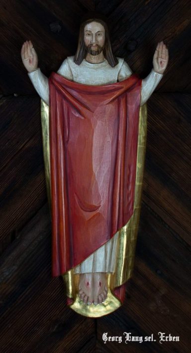 Christusfigur mit ausgebreiteten Armen, rot-goldenem Gewand, vor dunklem Hintergrund.