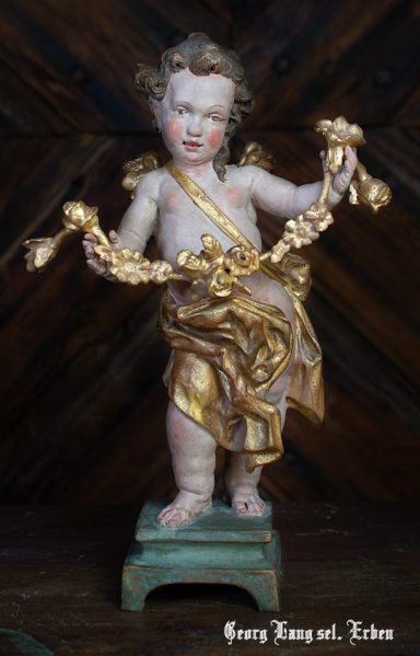 Ein kleiner Engel mit einer goldenen Girlande in der Hand, auf einem Sockel stehend.