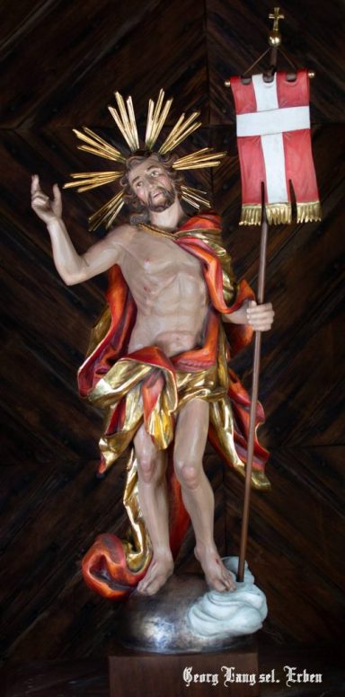 Christusstatue mit rotem Gewand, goldenen Strahlen und Fahne in der Hand.