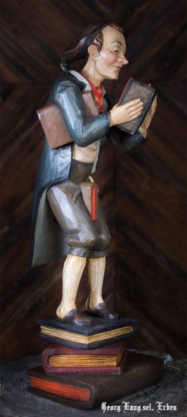 Eine geschnitzte Figur eines Mannes mit Bücherstapeln, der ein Buch hält.