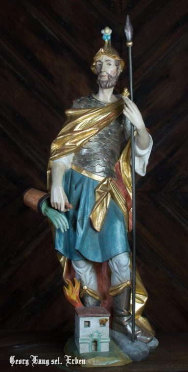 Heilige Figur in Rüstung mit Speer und Schwert, steht vor einem brennenden Haus.